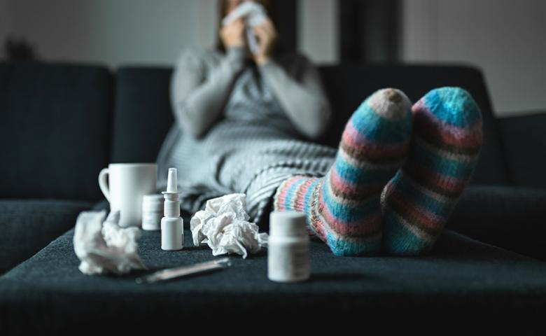A sick woman sits on a couch surrounded by tissues and medicine