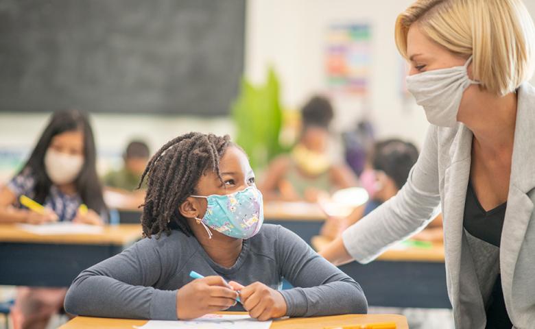 Female teacher leaning over a young female student&#039;s desk. Both wear face masks.