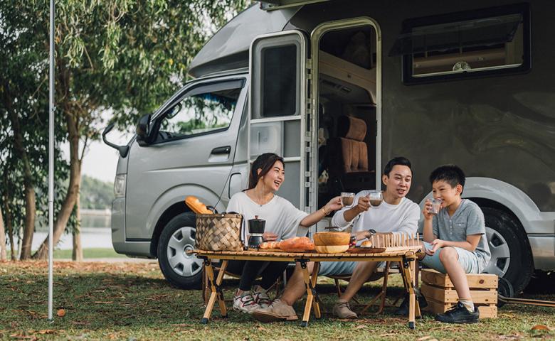 A young Asian family of three sits on the grass outside their camper van having a picnic. A lake is just visible in the back of the photo.