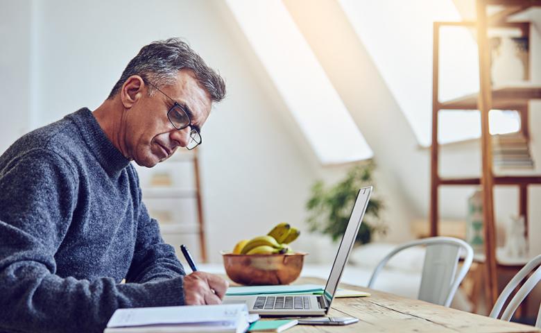 Man writing on notepad with laptop open.