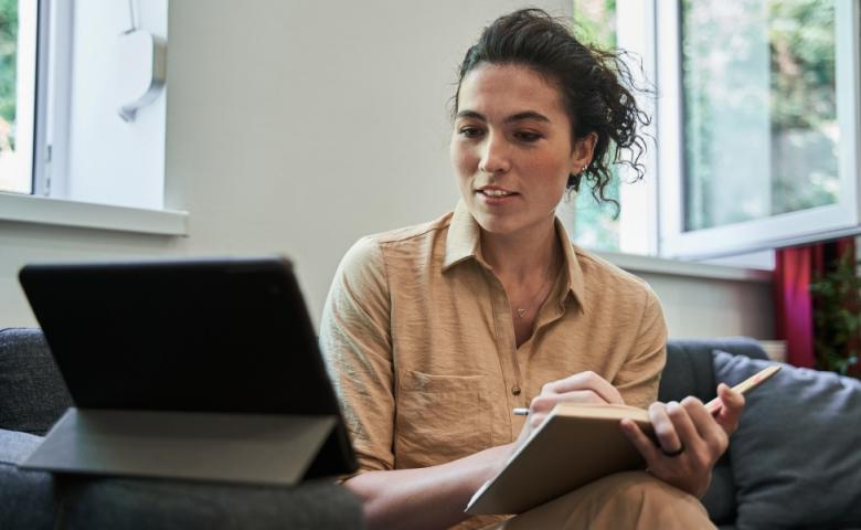 Person sitting on a couch taking notes in a notebook, with a tablet resting on the couch&#039;s arm. They appear to be watching something on the tablet&#039;s screen.