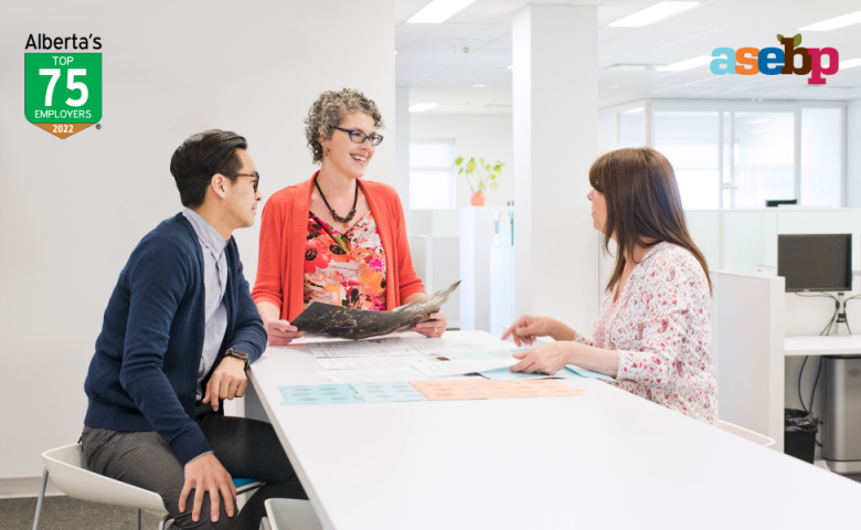 Photo of three employees working together at one of the collaboration station tables in ASEBP’s bright, open-concept office, pre-pandemic. Image includes Alberta’s Top Employers 2022 and ASEBP logos in the top corners.