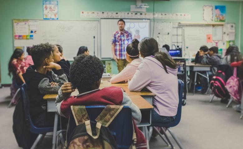 Male teacher stands at the front of a classroom, speaking to students.